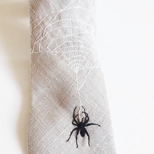 Eine beige Krawatte mit einem weißen gestickten Spinnennetz und einer schwarzen gestickten Spinne, die am Netz hängt.