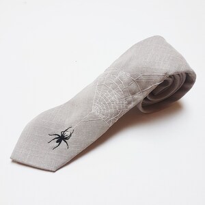 Eine graue Krawatte aus Leinen mit einem weißen gestickten Spinnennetz und einer schwarzen gestickten Spinne.