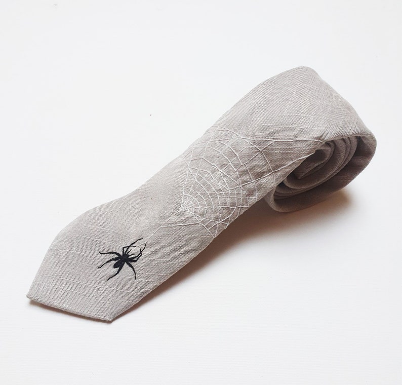 Eine graue Krawatte aus Leinen mit einem weißen gestickten Spinnennetz und einer schwarzen gestickten Spinne.