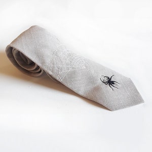 Eine graue Krawatte aus Leinen mit einem weißen gestickten Spinnennetz und einer schwarzen gestickten Spinne.