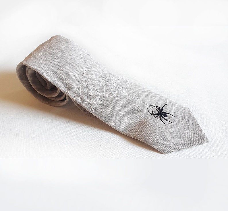 Eine graue Krawatte aus Leinen mit einem weißen gestickten Spinnennetz und einer schwarzen gestickten Spinne.