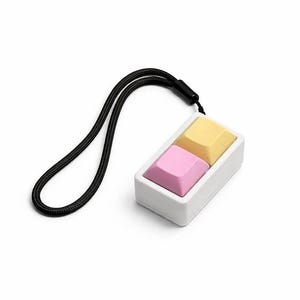 May include: A white rectangular pendant featuring two keyboard keycaps, one yellow and one pink. A black lanyard is attached to the pendant. The keycaps are set in a white frame.