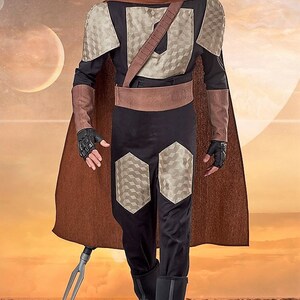 Pu&ograve; includere: Una persona che indossa un costume marrone e nero con un mantello marrone e una lancia d'argento. Il costume ha un motivo geometrico e una cintura marrone. La persona &egrave; in piedi in un ambiente desertico.