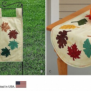 May include: A cream-colored garden flag featuring a circular wreath of multicolored fabric leaves and a matching table runner with scattered leaf appliques in shades of red, orange, brown, and green, accented by a decorative orange pumpkin.