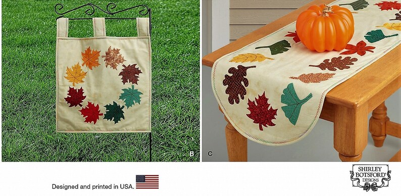 May include: A cream-colored garden flag featuring a circular wreath of multicolored fabric leaves and a matching table runner with scattered leaf appliques in shades of red, orange, brown, and green, accented by a decorative orange pumpkin.