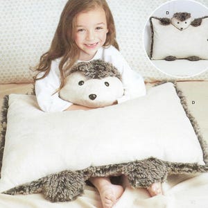 May include: A rectangular cream-colored pillow shaped like a hedgehog with a plush, dark brown faux fur border, a soft cream-colored center, and a detailed hedgehog head featuring dark eyes and a nose attached to the top edge of the pillow.
