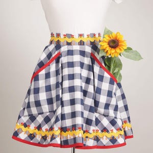 May include: A navy blue and white gingham apron featuring a yellow rickrack trim with embroidered chicken motifs along the waistband and hem. The apron includes two large pockets with red trim and a single yellow sunflower tucked inside one pocket.
