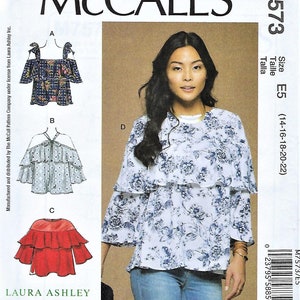 May include: McCall's sewing pattern M7573 featuring a woman wearing a floral print blouse. The pattern includes designs for several blouses with different styles, including off-the-shoulder and tiered ruffle details. The pattern is size E5 (14-16-18-20-22).