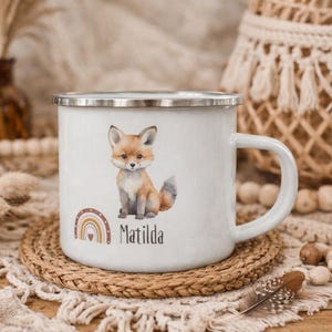 Puede incluir: Taza de esmalte blanco con una ilustración de acuarela de un zorro con pelaje marrón y gris. Un arcoíris y el nombre "Matilda" están debajo. La taza tiene un borde plateado y un asa blanca. Está sobre un posavasos tejido.