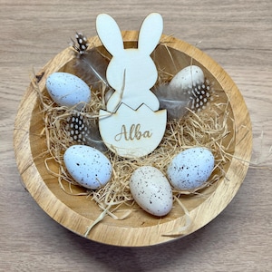 Peut inclure: Décoration de Pâques en bois avec un lapin sortant d'un œuf, gravé du nom "Alba". Le tout est dans un bol en bois contenant des œufs tachetés, de la paille et des plumes. Le bol est en bois clair.