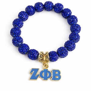 Puede incluir: Una pulsera de cuentas azules con un dije dorado. La pulsera presenta múltiples cuentas redondas azules brillantes. Un dije dorado con las letras ZΦB en azul cuelga de la pulsera. La pulsera está sobre un fondo blanco.