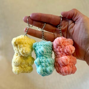 Puede incluir: Tres llaveros de oso de peluche en amarillo, turquesa y rosa. Cada oso está hecho de un material suave y texturizado y tiene un llavero plateado. Los osos son pequeños y tienen una forma redondeada.