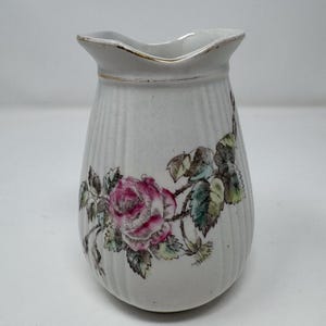 May include: A white ceramic pitcher featuring a vertical ribbed texture and a floral motif of a pink rose with green leaves. The rim of the pitcher is accented with a thin gold-colored trim, and the body has a slightly tapered, rounded shape.