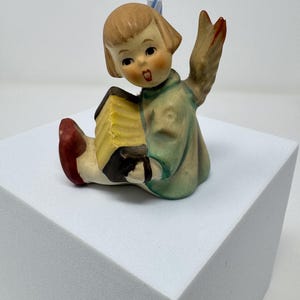 May include: A vintage ceramic figurine of a child angel playing an accordion. The angel wears a light green robe and has a red-tipped wing raised. The accordion is yellow and black. A blue candle is behind the angel's head.