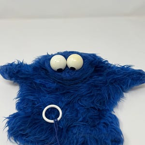 May include: A blue fuzzy hand puppet featuring two large white spherical eyes with black pupils. The puppet has a shaggy texture and includes a thin blue string attached to a white plastic ring hanging from the lower front section of the body.