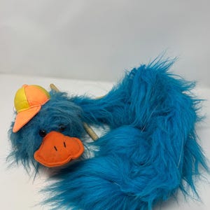 May include: A plush puppet featuring long, shaggy blue fur, a bright orange beak, and a yellow and orange baseball cap. The puppet has two small black eyes and a long, flexible body covered in soft, textured blue material.