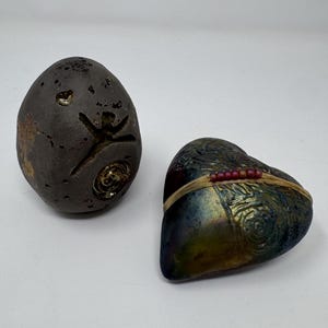 May include: A dark brown, textured egg-shaped object with carved symbols and a heart-shaped object with a metallic, iridescent finish, featuring a band of light-colored material adorned with a row of small, dark red beads across its surface.