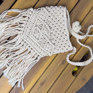 May include: Cream-colored macrame bag with a geometric design and long fringe. The bag has a braided strap and a knotted closure. The bag is made of a soft, woven material and is displayed on a wooden table.