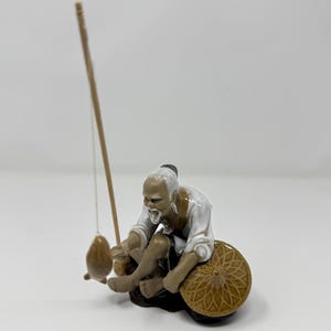 May include: A ceramic figurine of an elderly person with a white beard and hair, wearing a white shirt and brown pants, sitting while holding a tall fishing rod with a small brown fish attached to the line and a conical hat resting nearby.