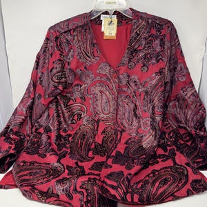 May include: A red paisley print blouse with a V-neck and long sleeves. The blouse has a button closure and is hanging on a hanger. The paisley pattern includes black and gold accents.