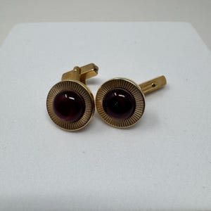 May include: A pair of gold-tone cufflinks featuring a circular design with a deep burgundy cabochon center. The outer edge of each cufflink has a textured, sunburst-style radial pattern surrounding the smooth, dark red stone inlay.