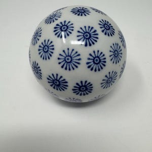 May include: A spherical ceramic object with a glossy white finish, decorated with a repeating pattern of small, dark blue starburst or floral motifs distributed evenly across its entire rounded surface.
