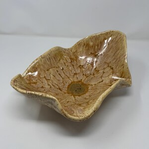 May include: A ceramic bowl with an irregular, wavy rim and a glossy finish. The surface features a mottled pattern of tan, beige, and cream tones, radiating from a darker, speckled center. The bowl has a fluid, organic shape with raised edges.