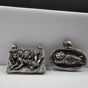 May include: Two silver-toned metal pendants. The rectangular pendant features a relief of a winged fairy figure. The oval pendant displays a relief of a cat with a textured body. Both pieces have a loop at the top for attachment to a chain or cord.