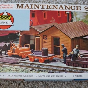 May include: A model train maintenance shed kit with a wooden structure, figures, and vehicles. The box features the text "MAINTENANCE SHED" and "H-O scale realism!" Includes a motor car and trailer. The kit is in realistic colors.