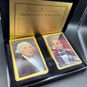 May include: A black box set containing a deck of playing cards featuring portraits of U.S. Presidents. The cards have gold edges and are displayed in a velvet-lined case. The box includes a gold plaque with text.