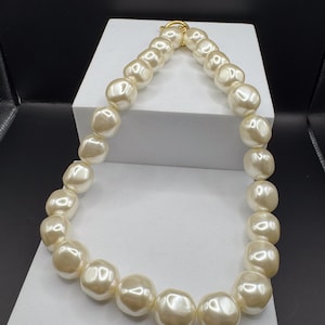 May include: A necklace composed of large, off-white, irregularly shaped faux pearl beads with a glossy finish. The beads are strung together in a single strand, connected at the top by a circular gold-toned metal toggle clasp mechanism.