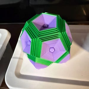 May include: A geometric, multi-faceted sculpture in shades of green and lavender. The object is a complex arrangement of layered shapes, creating a visually interesting three-dimensional form. It sits on a white tray.