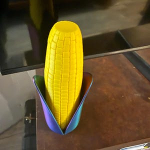 Puede incluir: Un objeto amarillo en forma de mazorca de maíz con una superficie texturizada, colocado en un soporte iridiscente multicolor. El soporte tiene forma de tulipán con tonos azules, morados y verdes. El artículo está sobre una superficie de madera marrón.