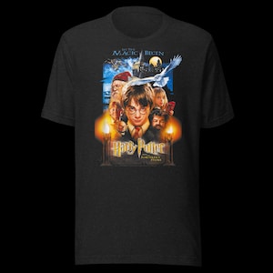 Op de afbeelding: Een antracietgrijs T-shirt met korte mouwen en een grafische print van de filmposter Harry Potter en de Steen der Wijzen. Het ontwerp bevat de tekst Let The Magic Begin, de titel en illustraties van de hoofdpersonages.