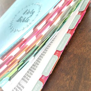 Puede incluir: Una Biblia abierta con pestañas de página coloridas. La portada es azul claro con las palabras "Holy Bible" en relieve. Las pestañas son de varios colores, incluyendo rosa, verde y rojo, con nombres de capítulos visibles.