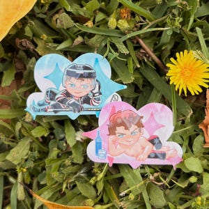 Op de afbeelding: Twee cartoon-stijl pins met mannelijke figuren. Eén is gekleed in hockey-uitrusting, de andere is shirtless. Beide staan op hartvormige achtergronden met steraccenten. De pins staan tegen een achtergrond van groen gras, bladeren en een gele paardenbloem.
