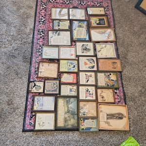 May include: A collection of framed vintage prints and illustrations, arranged on a patterned rug. The frames vary in size and style, with a mix of artwork including text, drawings, and colorful images. The rug has a dark background with a pink and red floral design.