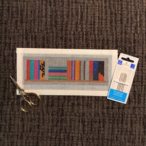 May include: A needlepoint canvas featuring a bookshelf design with colorful books. Gold scissors and a package of tapestry needles are next to the canvas. The canvas is on a brown textured surface.