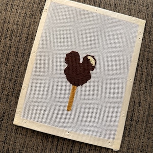 May include: A needlepoint canvas featuring a dark brown Mickey Mouse ice cream bar design. The ice cream is on a light brown stick, with a white and yellow accent. The canvas is rectangular with a white mesh and a beige border.