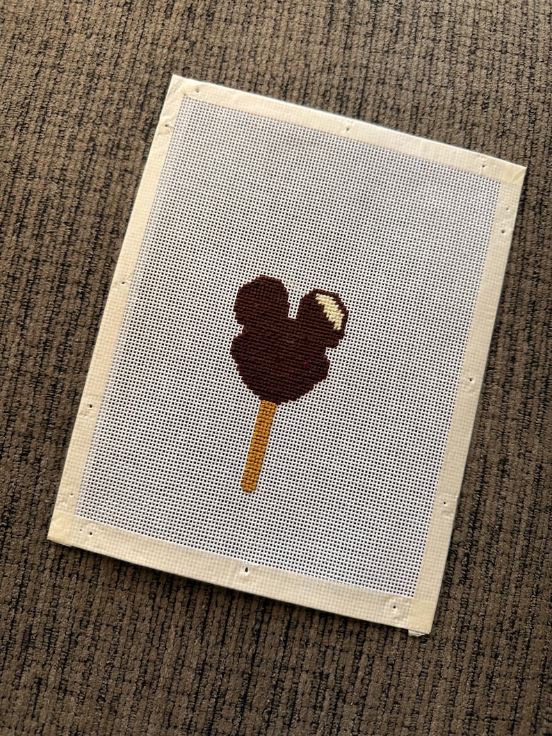 May include: A needlepoint canvas featuring a dark brown Mickey Mouse ice cream bar design. The ice cream is on a light brown stick, with a white and yellow accent. The canvas is rectangular with a white mesh and a beige border.