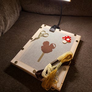 May include: A needlepoint project in progress, featuring a wooden frame with a canvas displaying a brown Mickey Mouse ice cream bar design. Gold scissors, a red mushroom pin, and skeins of yellow, brown, and cream-colored thread are present. A black LED lamp is attached.