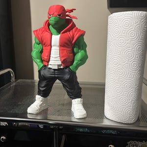 May include: A green anthropomorphic turtle figurine wearing a red headband, a red sleeveless hoodie over a white shirt, black pants, and white sneakers. The figure stands next to a white paper towel roll on a metallic surface with a perforated top.