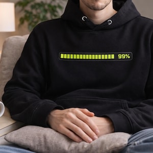 May include: Black hoodie featuring a neon green loading bar graphic with "99%". The hoodie has a drawstring hood. A person is wearing the hoodie while sitting on a sofa. A white mug of coffee sits on a stack of books.