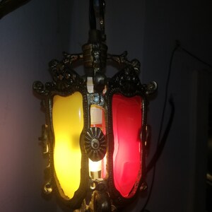 May include: Ornate, antique-style hanging lamp with a bronze-colored frame. The lamp features two colored glass panels, one yellow and one red, with decorative metalwork and a central light source. The lamp is suspended from a black cord.