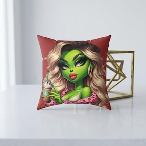 May include: A decorative square pillow with a cartoon image of a green-skinned character with blonde hair, holding a coffee drink. The character is wearing a pink leopard print top and gold hoop earrings. The pillow has a red background.