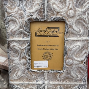 May include: A decorative, rectangular picture frame with a distressed, textured finish. The frame is made of cast wood and measures 5" x 7". The frame has a brown and white color scheme and features the text "Sixth Sister Custom Frames & More".