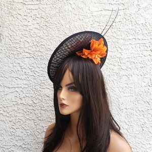 May include: A black mesh fascinator with an orange flower accent and two black feather accents. The hat is oval-shaped and sits atop a mannequin's head. The background is a textured white wall.