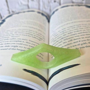 May include: A light green, diamond-shaped book page holder with a circular opening. The book holder is resting on the pages of an open book. The book's text is visible in the background.