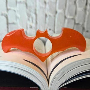 Puede incluir: Un sujetapáginas naranja con forma de murciélago hecho de resina. El soporte se coloca sobre un libro abierto, con las alas del murciélago extendiéndose sobre las páginas. Tiene una abertura circular en el centro.