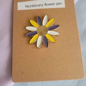 May include: A daisy flower pin with yellow, white, and purple petals, and a gold center. The pin is on a brown card with the text "Nonbinary flower pin". The background is a light purple and blue fabric.